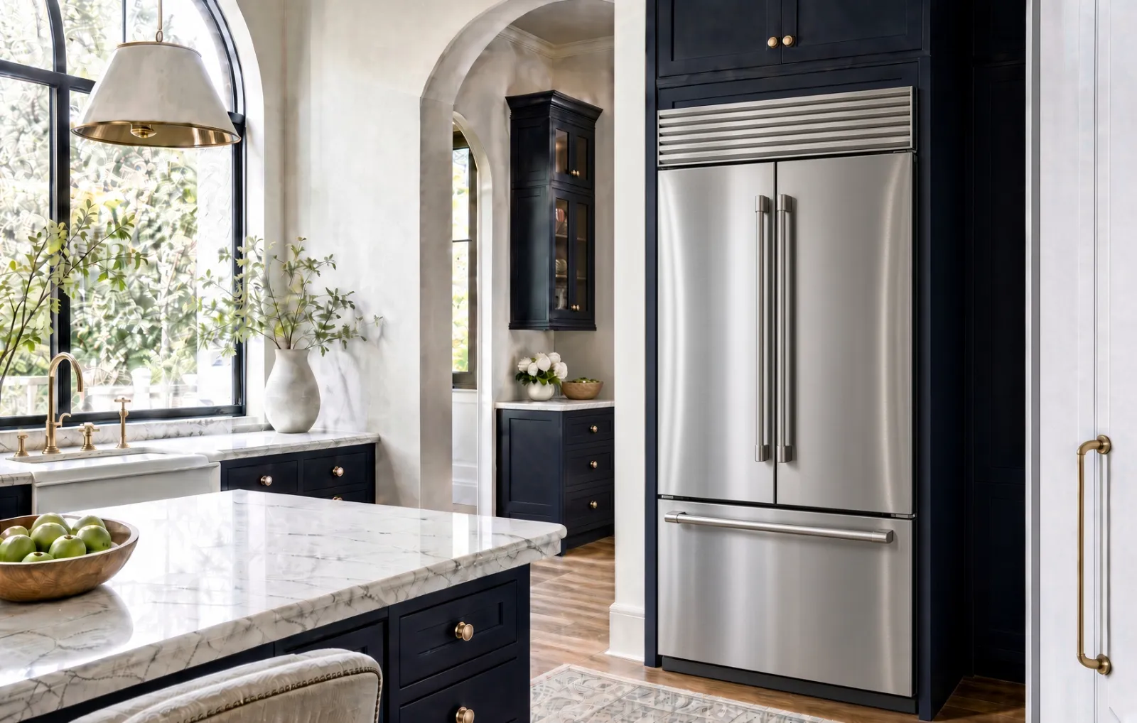 Stainless steel French-door built-in refrigerator with louvered top grille set into navy cabinetry beside a marble island
